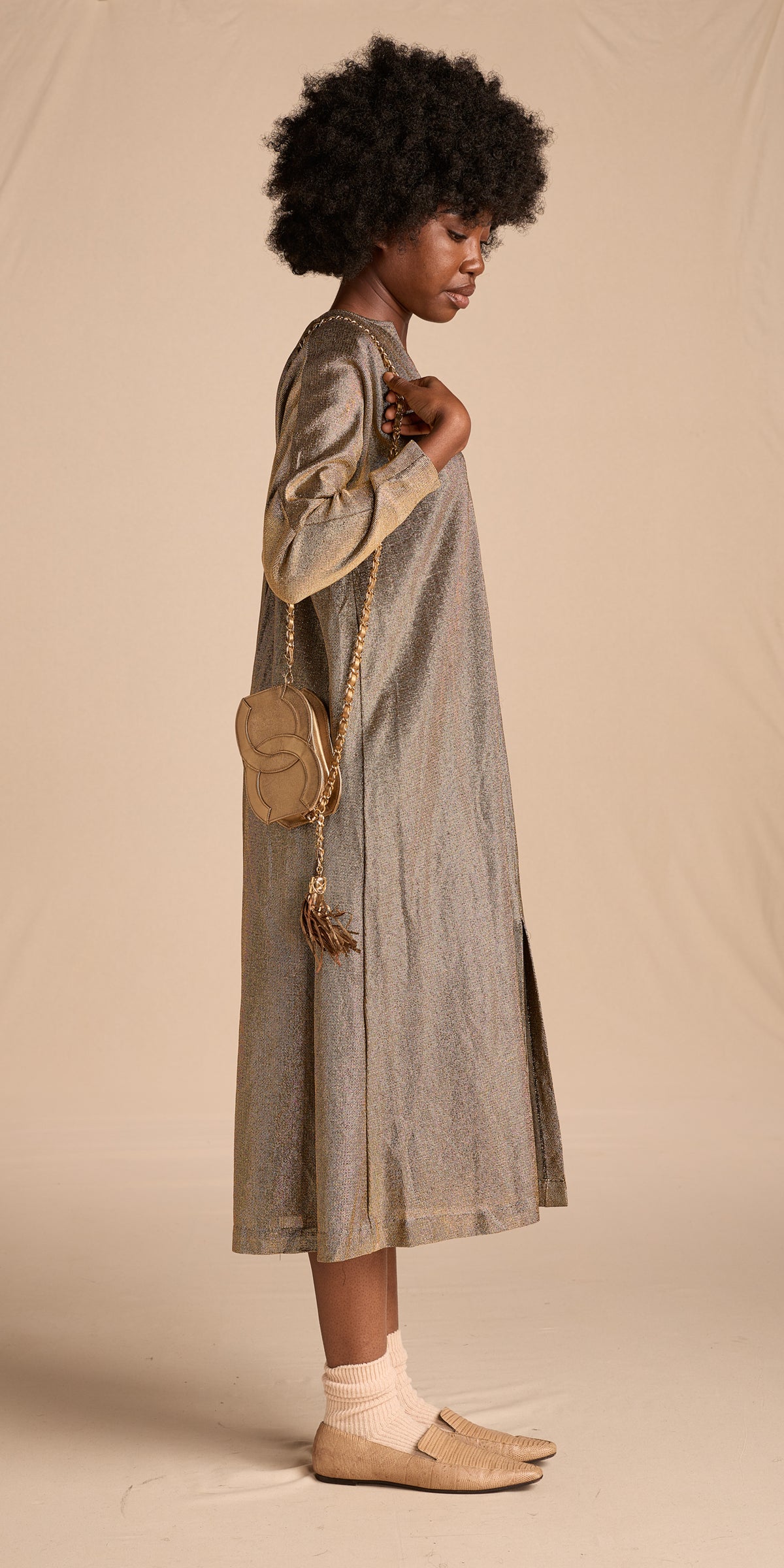A model wearing a long, loose-fitting gold dress with side slits, paired with a small woven shoulder bag, cream socks, and tan loafers, standing against a warm beige studio backdrop.