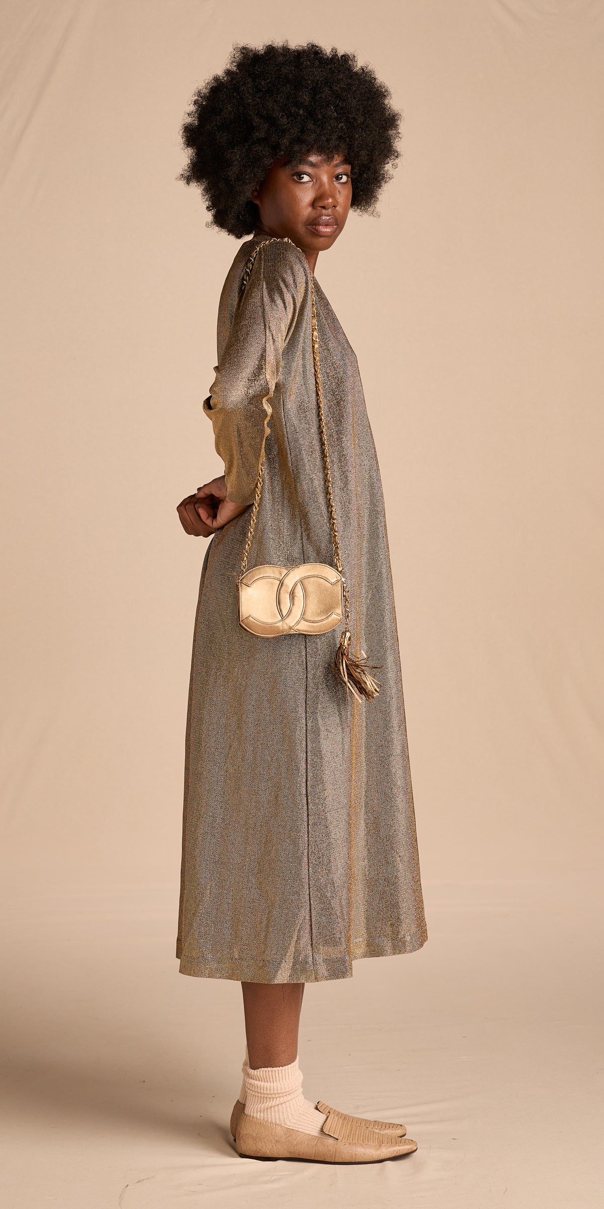 A model is shown from the back wearing a long-sleeved, knee-length metallic taupe dress, a small gold chain crossbody bag with a tassel, and beige loafers, set against a solid beige studio backdrop.