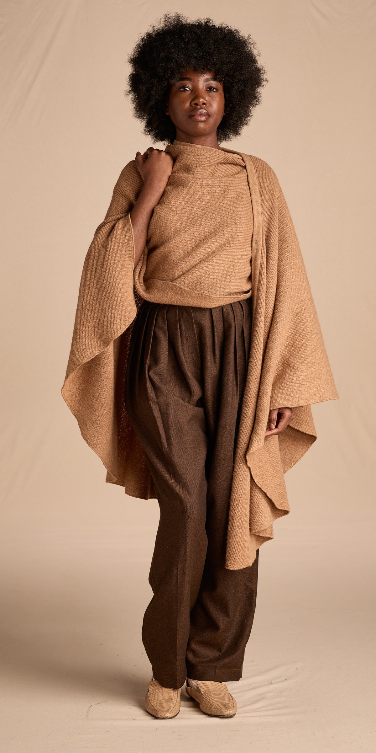 A model stands against a beige studio backdrop wearing a camel-colored knit poncho draped over a beige turtleneck, dark brown wide-leg pants, and tan mule shoes.