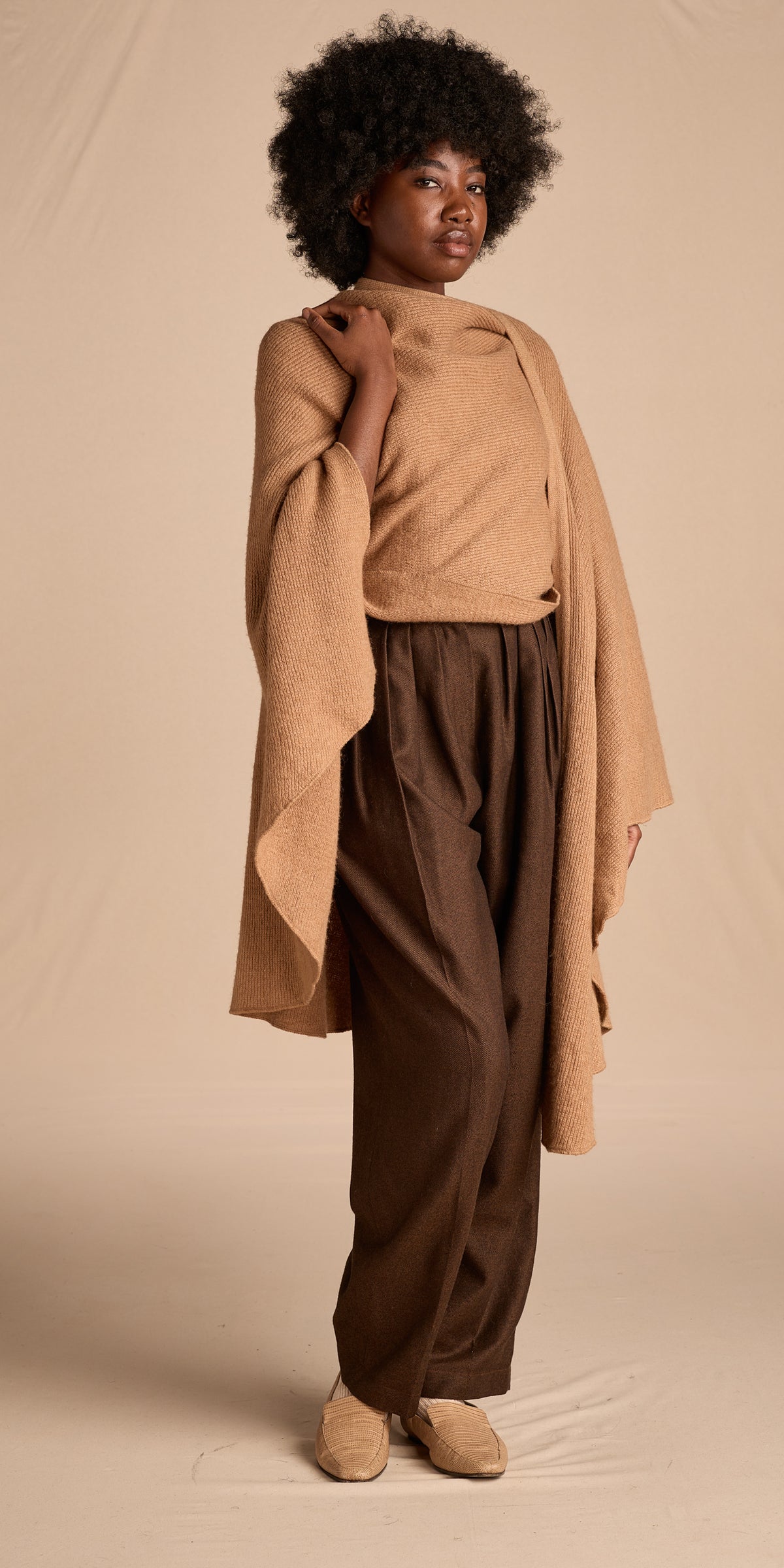 A model stands against a warm beige studio backdrop, wearing a camel-colored knit mock-neck top with draped sleeves, dark brown wide-leg trousers, and tan slip-on shoes.